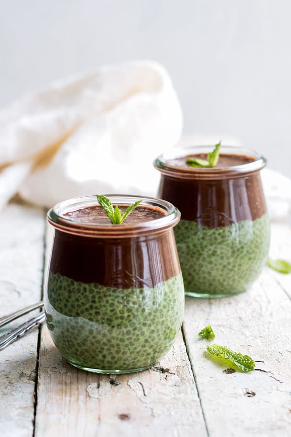 two jars of green chia pudding with chocolate smoothie on top on a white wooden table