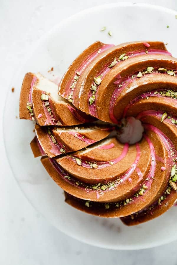 an orange cardamom bundt cake on a cake stand with pink blood orange glaze and chopped pistachios