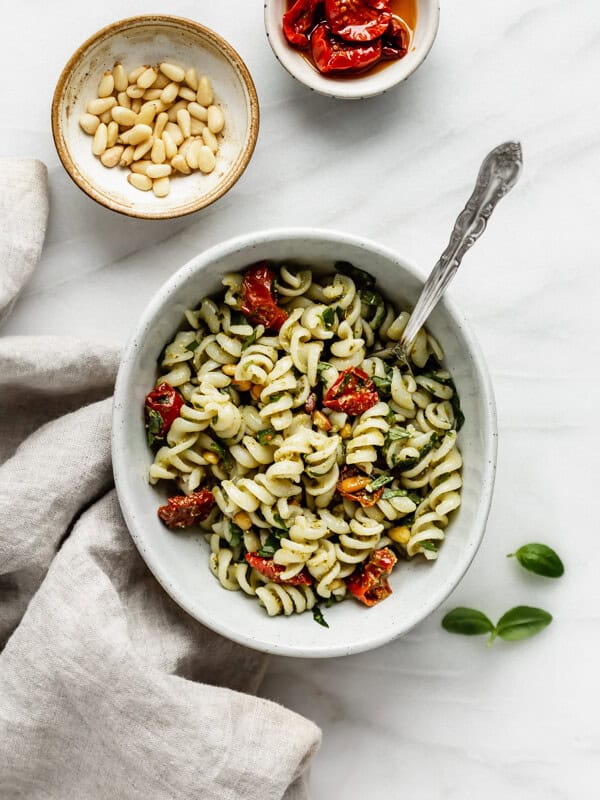 a bowl of pesto pasta salad with a napkin and some pine nuts on the side