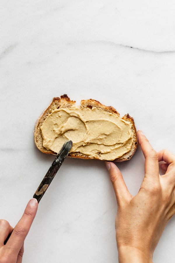 a hand holding a knife spreading hummus on toast