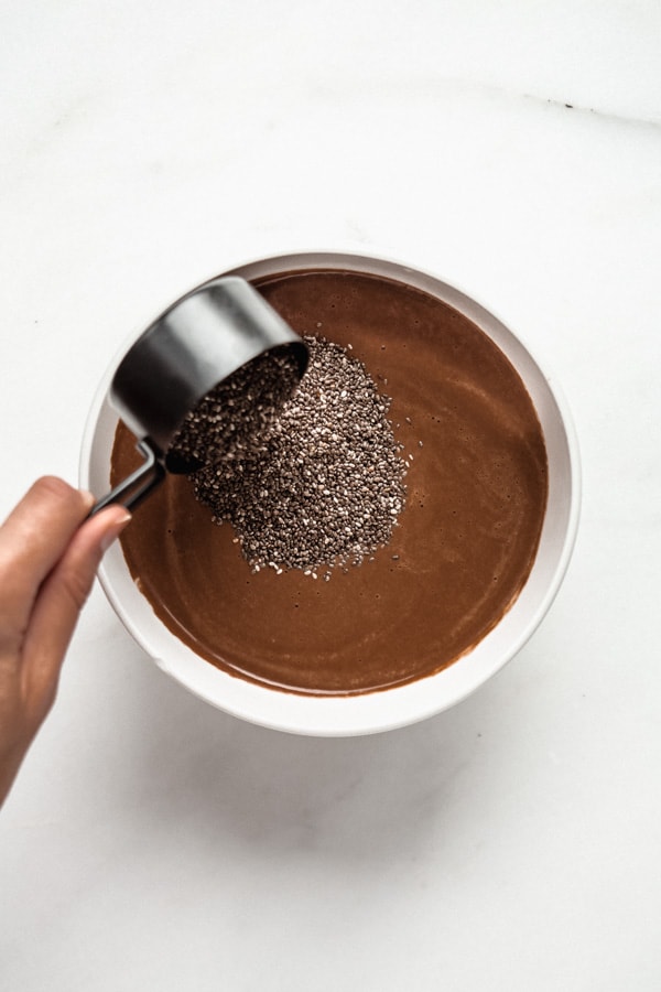 a measuring cup pouring chia seeds into a bowl of cashew chocolate milk