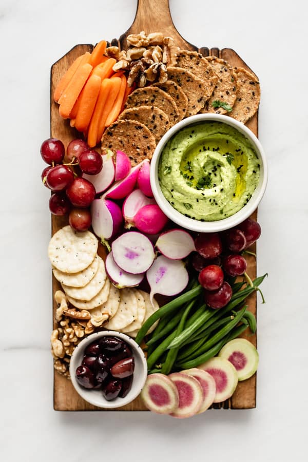 a wooden platter with veggies, fruits, crackers and edamame hummus on it