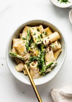 a bowl of lemon asparagus rigatoni noodles topped with small bowls of parsley and chilli flakes on the side