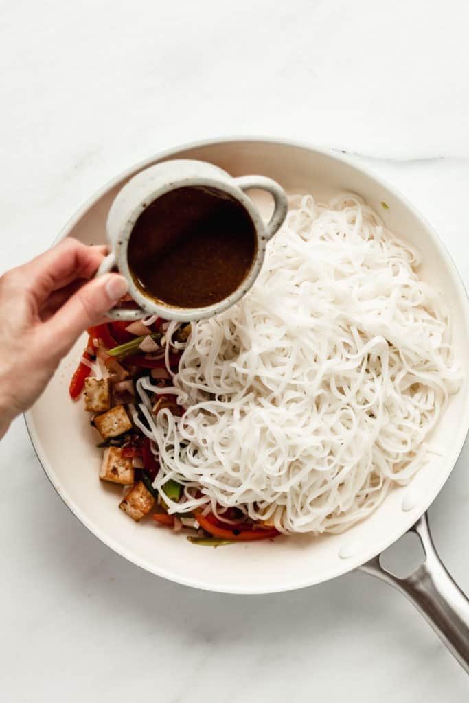rice noodles in a pan with a hand pouring sauce onto them
