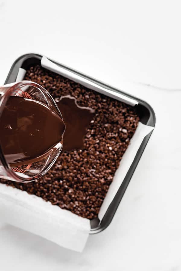 a measuring cup pouring chocolate into a pan of chocolate rice Krispie treats