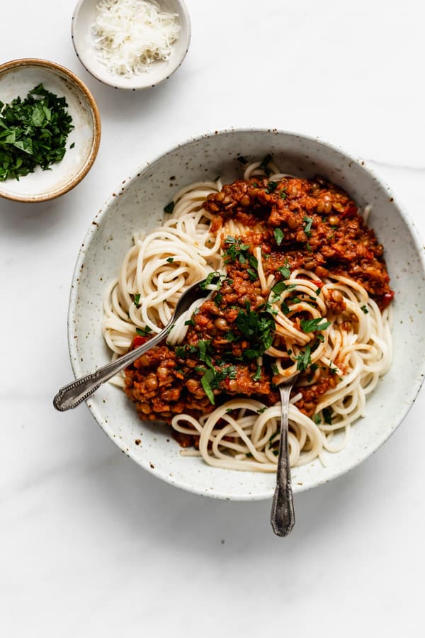 a white bowl filled with spaghetti and lentil bolognese and two smaller bowls on the side with parsley and parmesan cheese