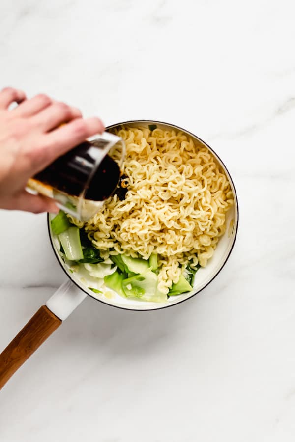 a hand pouring sauce onto ramen noodles in a white pot