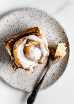 a cinnamon roll on a white speckled plate topped with cream cheese frosting