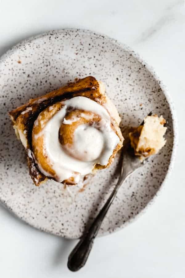 a cinnamon roll on a white speckled plate topped with cream cheese frosting