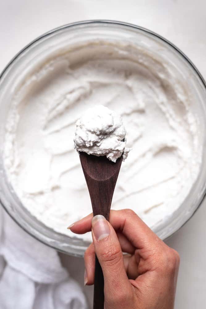 hand holding a wooden spoon of coconut yogurt over the large glass bowl of yogurt