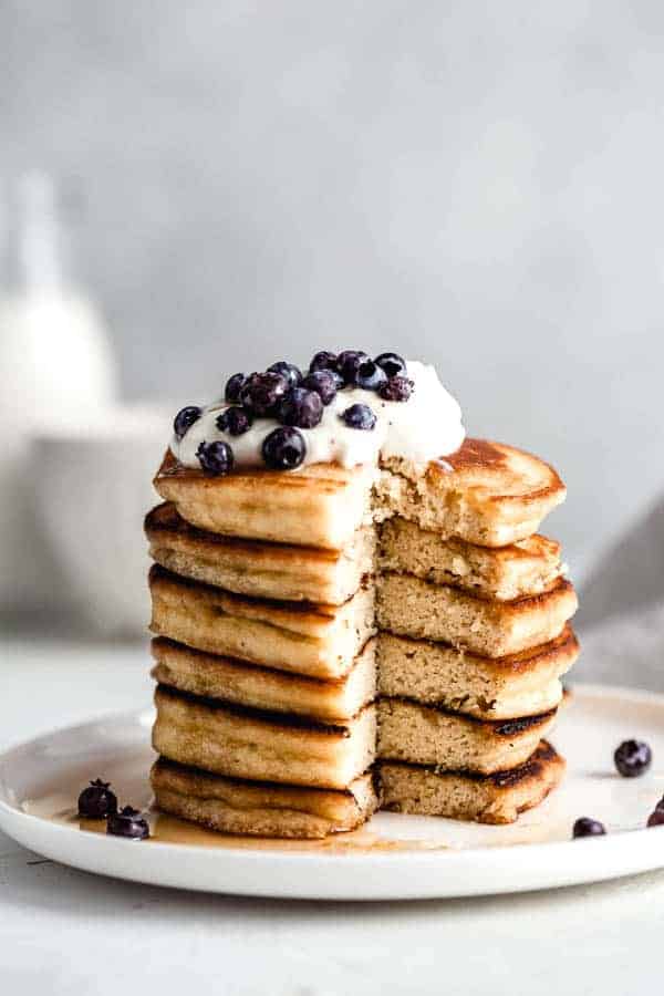 healthy breakfast pancakes topped with cream and blueberries, on a white plate against a grey background