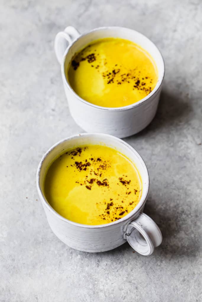 two white mugs filled with yellow turmeric latte and topped with cinnamon, on a grey background