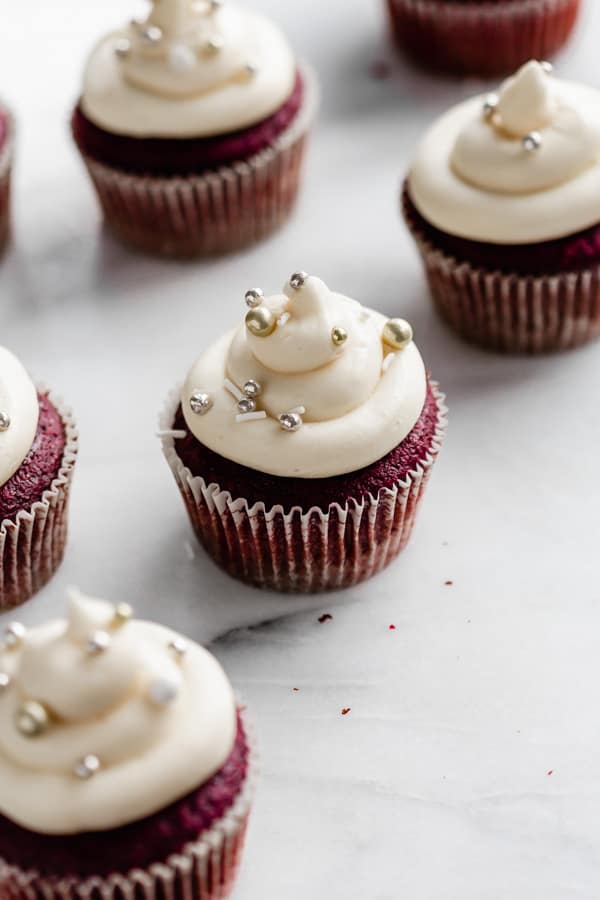 vegan red velvet cupcakes on a marble board topped with white frosting and silver ball sprinkles