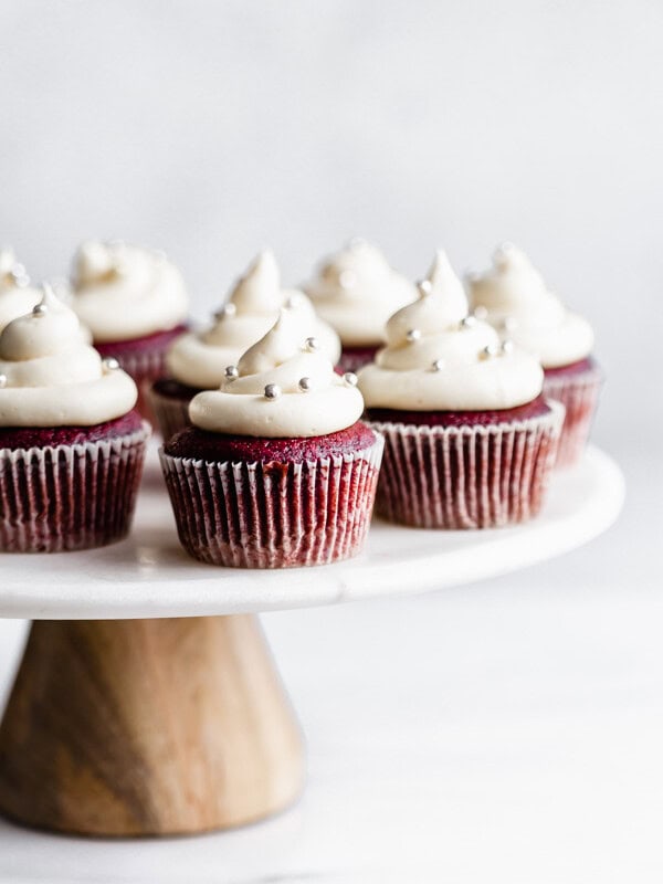 vegan red velvet cupcakes with cream cheese frosting and silver sprinkles on a marble and wood cakestand