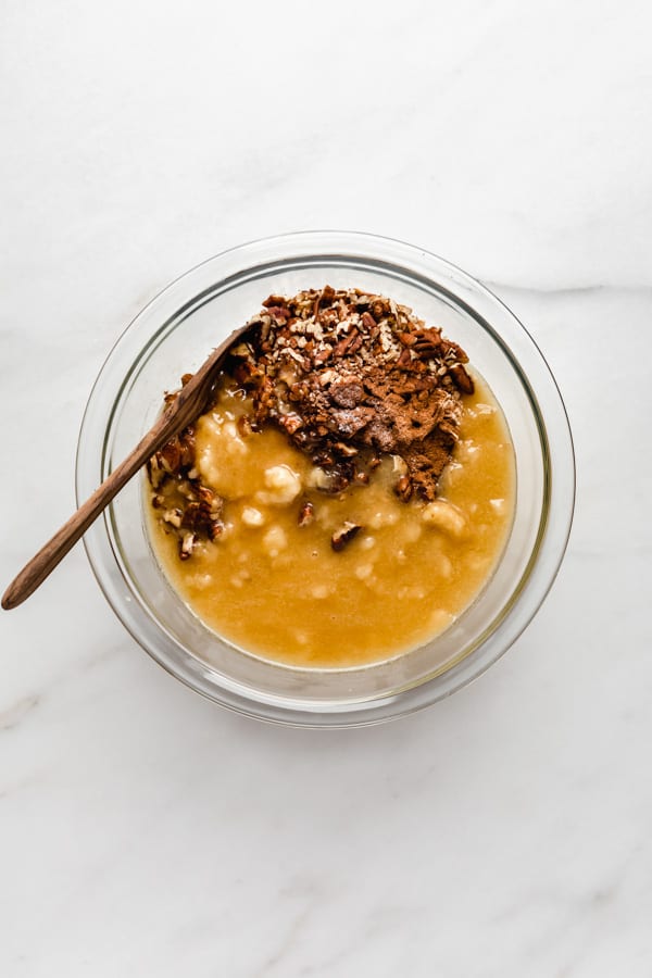 a clear mixing bowl with ingredients to make banana bread granola and a wooden spoon in it
