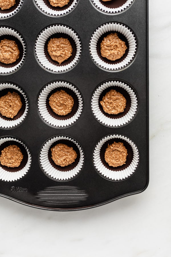 a muffin pan filled with muffin liners, a layer of chocolate and a scoop of almond butter