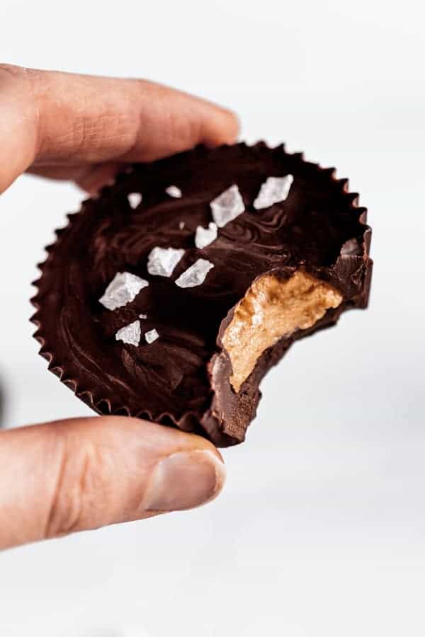 A hand holding a dark chocolate almond butter cup with a bite taken out of it