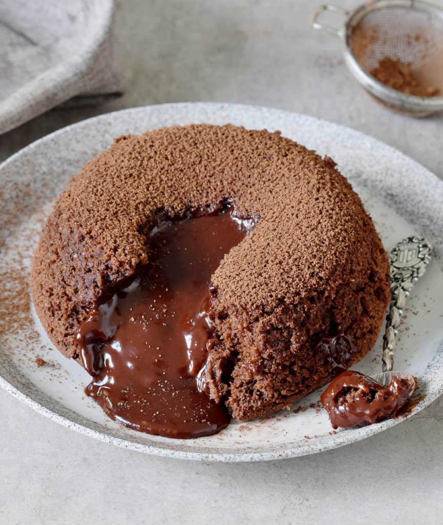 molten chocolate spilling out of a chocolate lava cake on a grey plate with a silver spoon covered in chocolate on the side