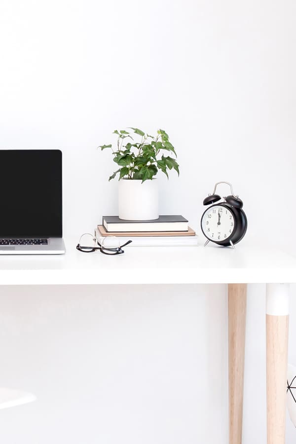 A clock, stack of books and plant on a white desk