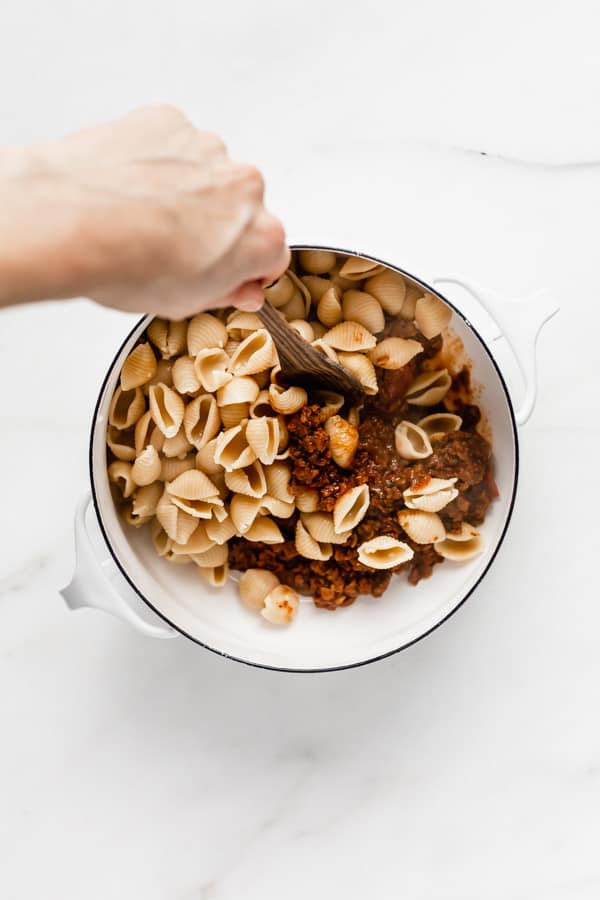 a hand mixing vegan taco sauce with shell noodles in a white pot