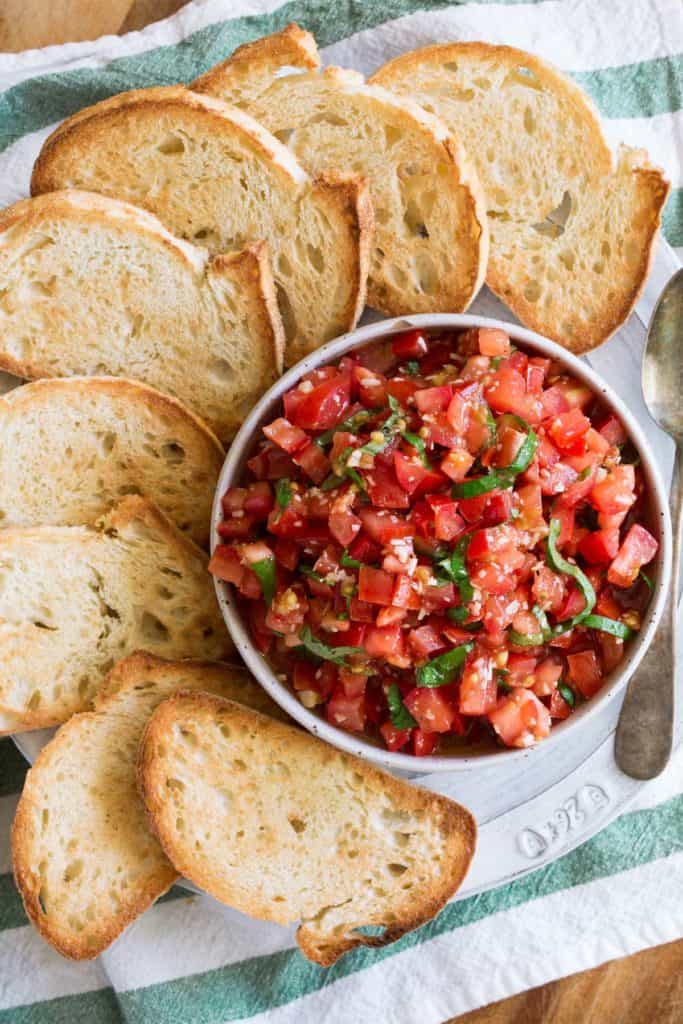 tomato bruschetta in a white bowl surrounded by toasted bread against a green and white linen