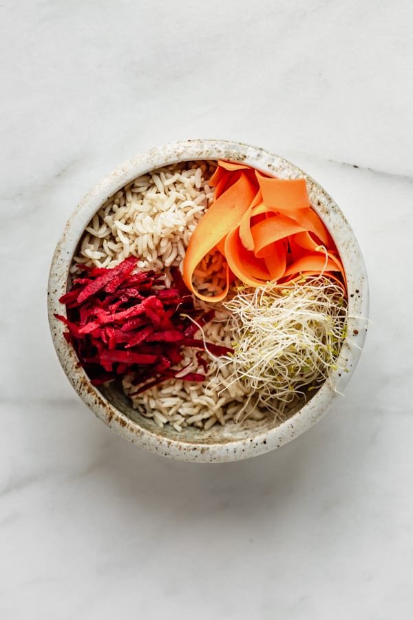 a white ceramic bowl with brown rice, beets, carrots and sprouts in it