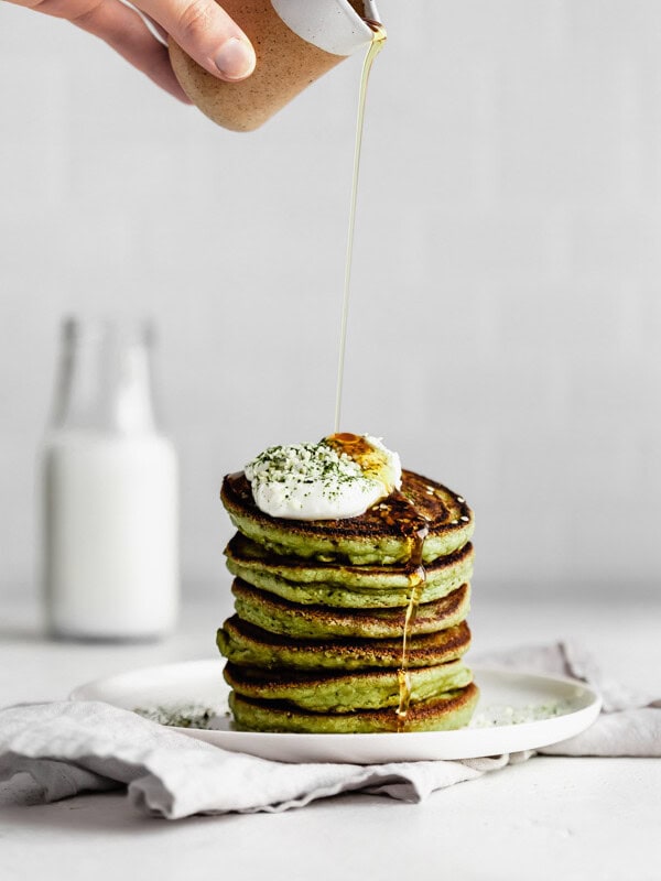 a hand pouring syrup onto a stack of matcha pancakes