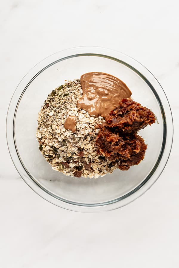 A clear mixing bowl with oats, nuts, date paste and almond butter in it