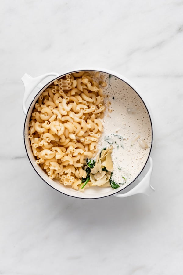 A white pot with spinach, artichoke hearts, vegan cream sauce and macaroni noodles in it