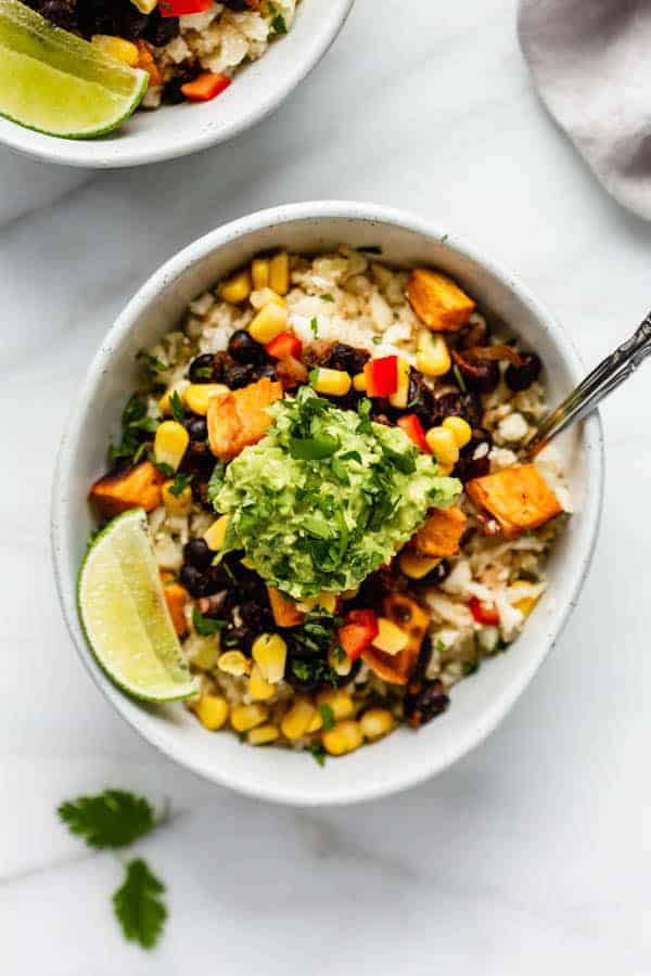 burrito bowl in a white bowl with corn, black beans, peppers, and a sliced lime topped with guacamole and chopped cilantro