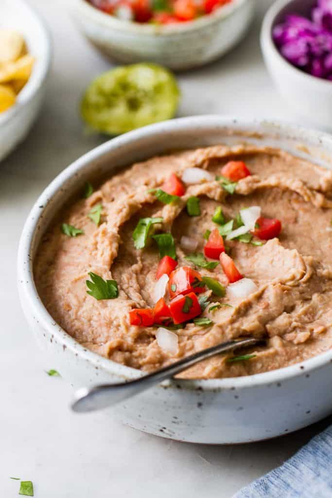 white speckled ceramic bowl filled with refried beans topped with fresh tomatoes and onions with small bowls of ingredients in the background