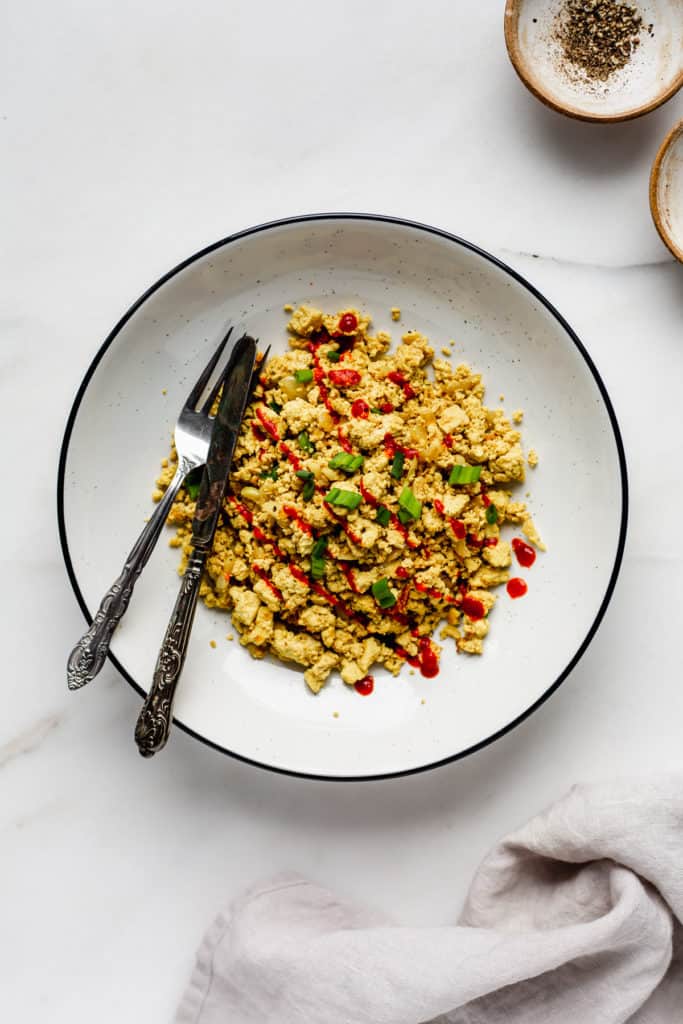 A bowl of scrambled tofu with a fork and knife in it and a small pinch bowl of ground pepper on the side
