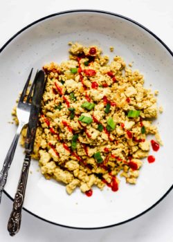 scrambled tofu topped with sliced green onions and sriracha in a large white bowl with a black rim