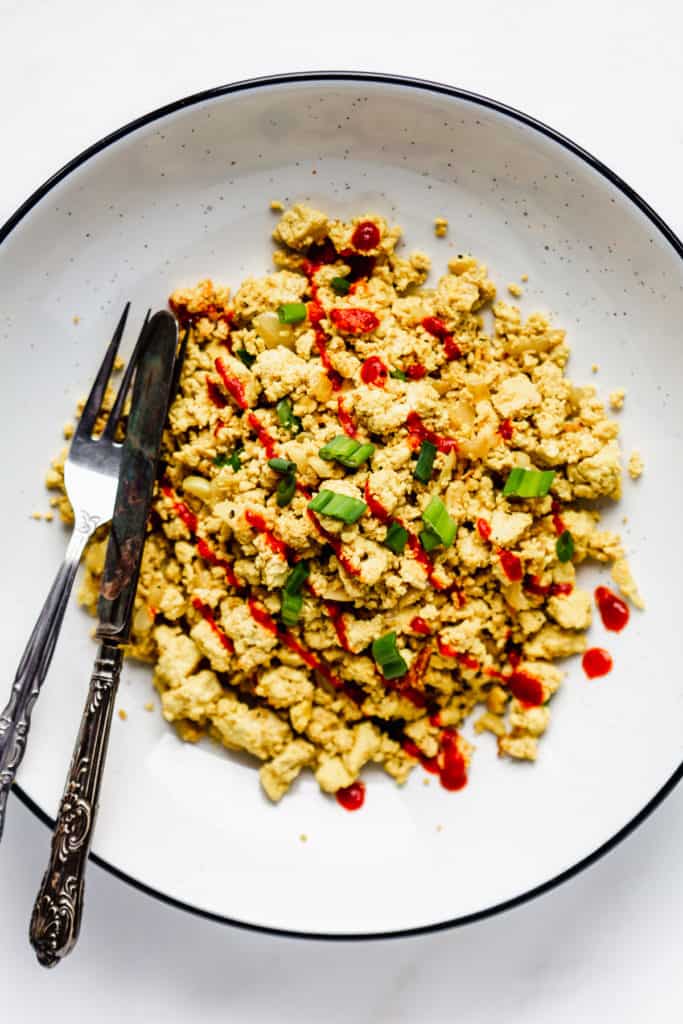 scrambled tofu topped with sliced green onions and sriracha in a large white bowl with a black rim