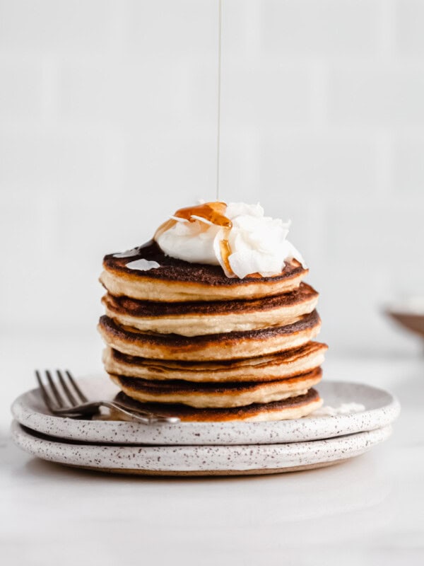 a stack of coconut flour pancakes topped with yogurt and coconut flakes with maple syrup being drizzled on top