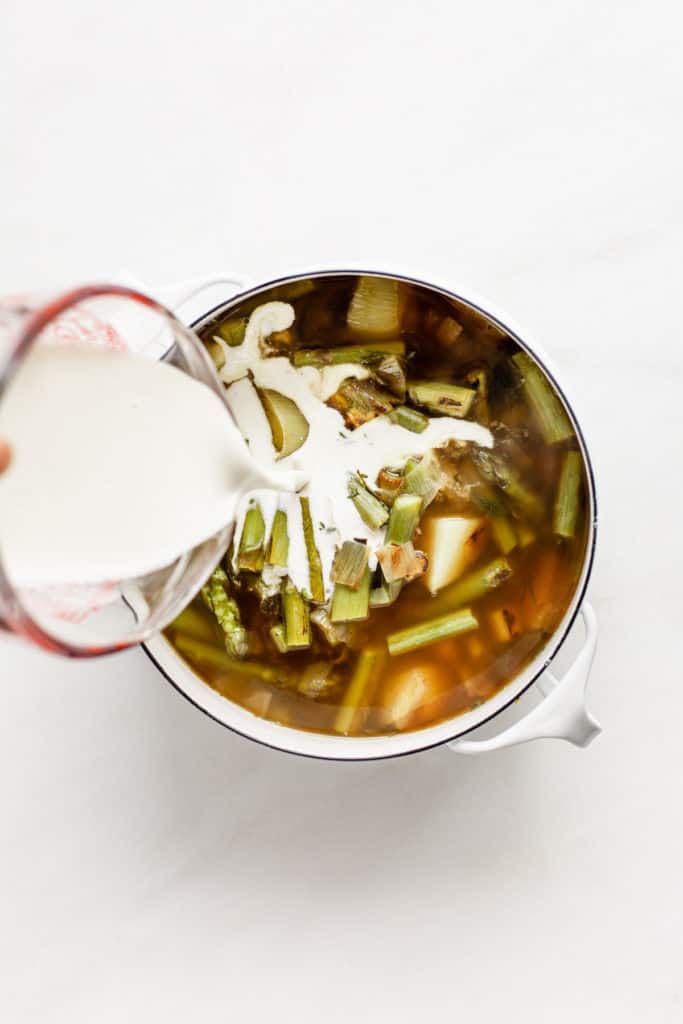 A measuring cup pouring cashew cream into a pot of broth with asparagus and vegetables