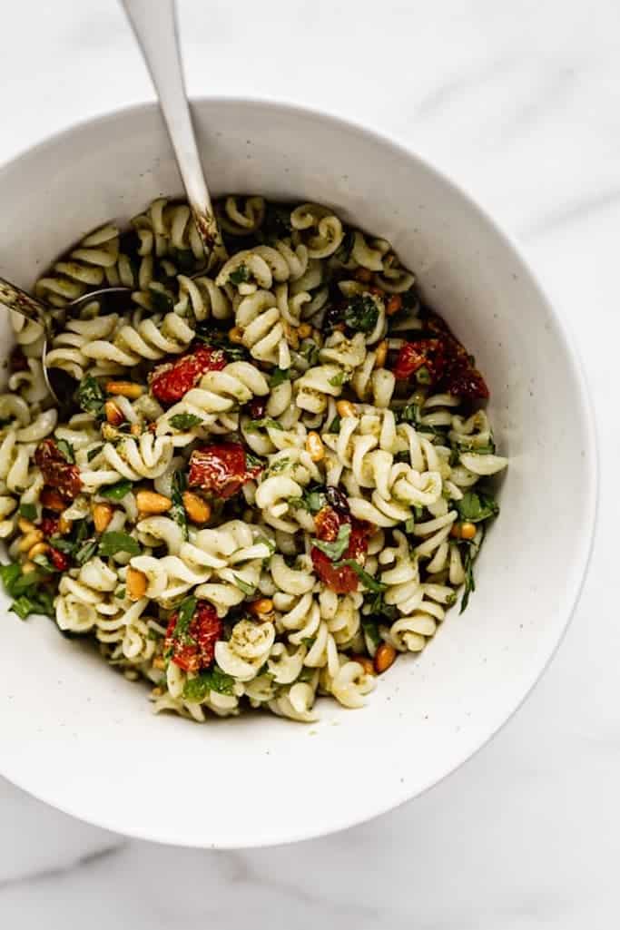 fusilli pasta covered in pesto mixed with sundried tomatoes and pine nuts in a white bowl with a silver spoon