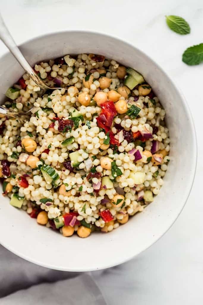 white serving bowl filled with israeli couscous mixed with fresh vegetables and herbs and a silver spoon