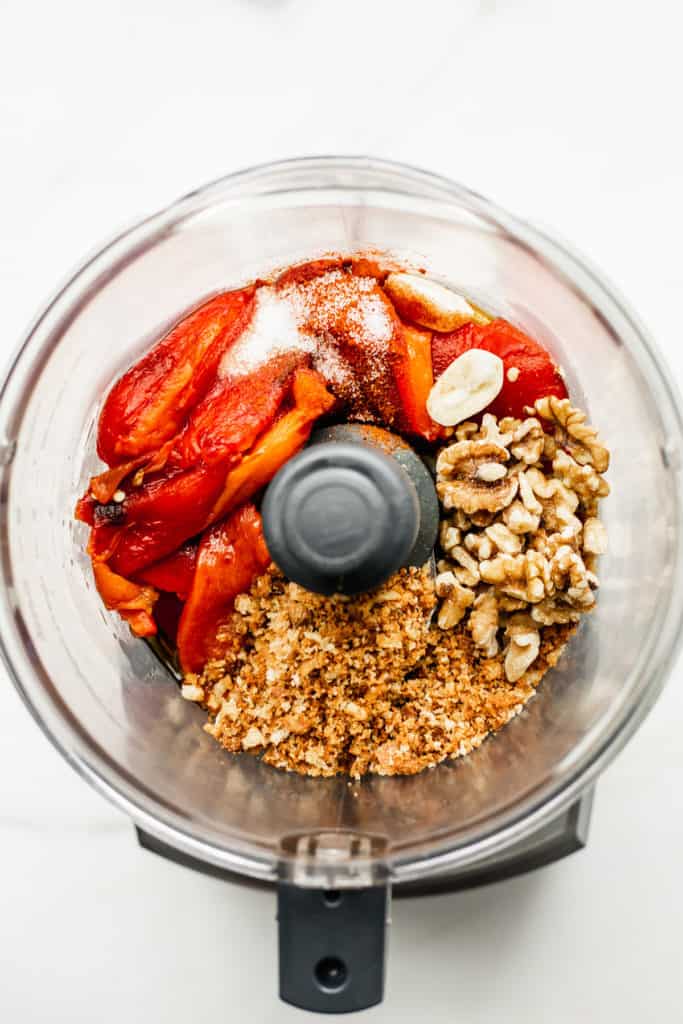 a food processor with red peppers, breadcrumbs, walnuts and garlic in it