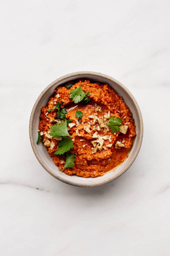 a ceramic white bowl with roasted red pepper dip topped with crushed walnuts and parsley