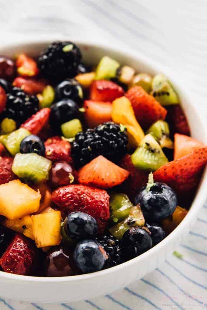 white bowl of fruity summer salads of blackberries, strawberries, kiwi, and mango against a white and blue striped linen