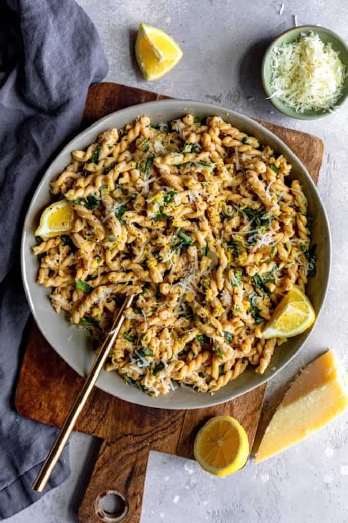 twisted pasta noodles covered in lemon basil sauce with shredded asiago cheese and lemon slices on a grey backdrop, summer salads with lemon wedges