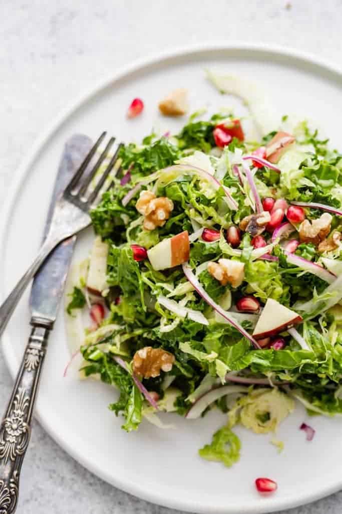 summer salads made with shaved brussels sprouts, kale, fennel, and chopped apples topped with walnuts and pomegranate seeds on a glossy white plate with a silver fork and knife