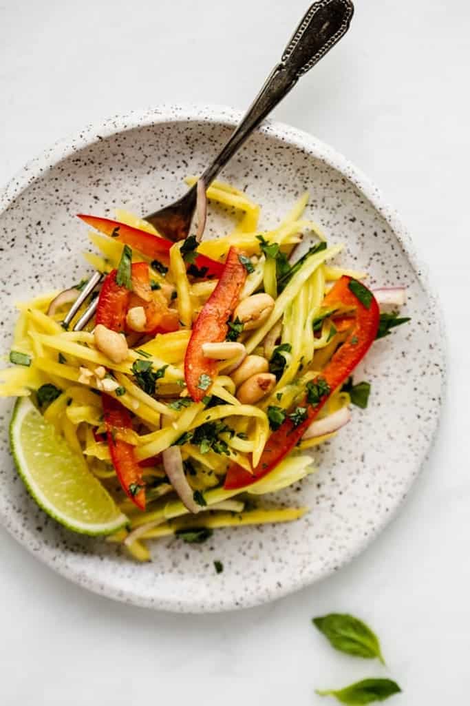 thinly sliced mango, red onion, and red pepper topped with roasted peanuts on a white speckled plate with a silver fork on a white marble table