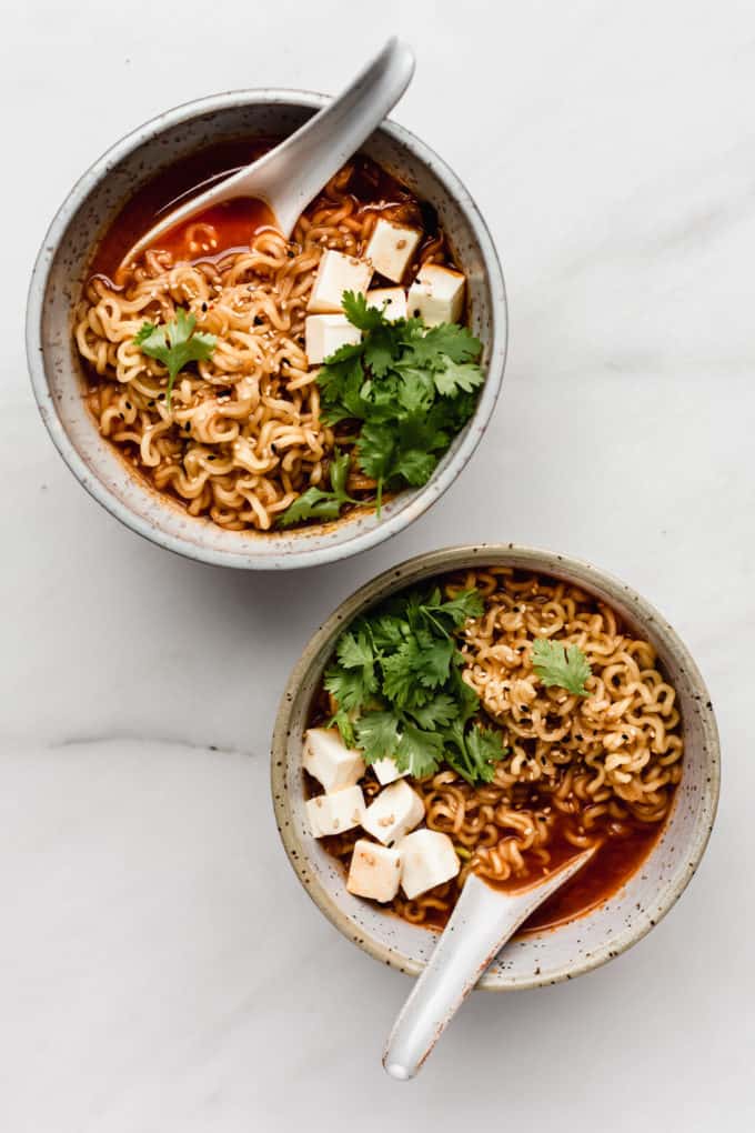 two white speckeld ceramic bowls of kimchi ramen with spoons in them