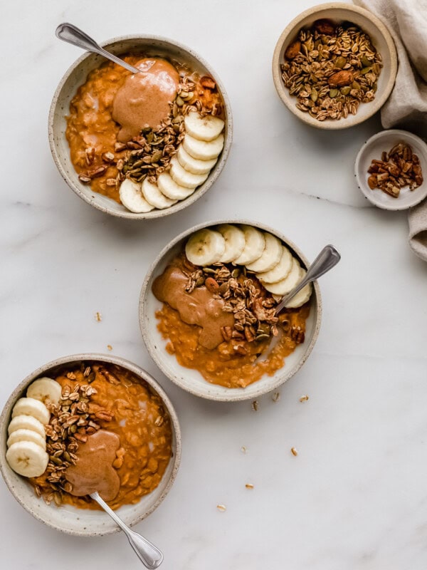 three bowls of pumpkin pie oatmeal with small bowls of granola and pecans on the side