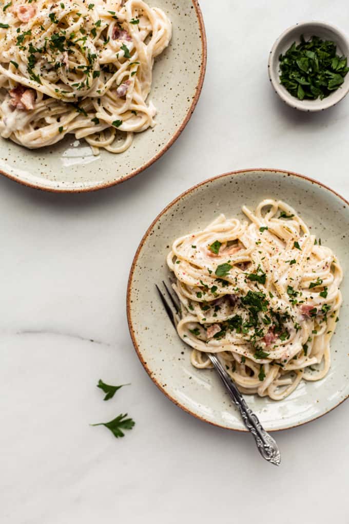 Two plates of vegan carbonara with a small bowl of parsley on the side