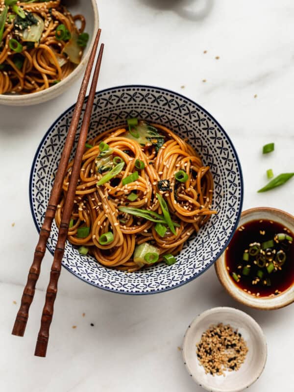 Teriyaki noodles in a blue bowl with a side of teriyaki sauce and sesame seeds