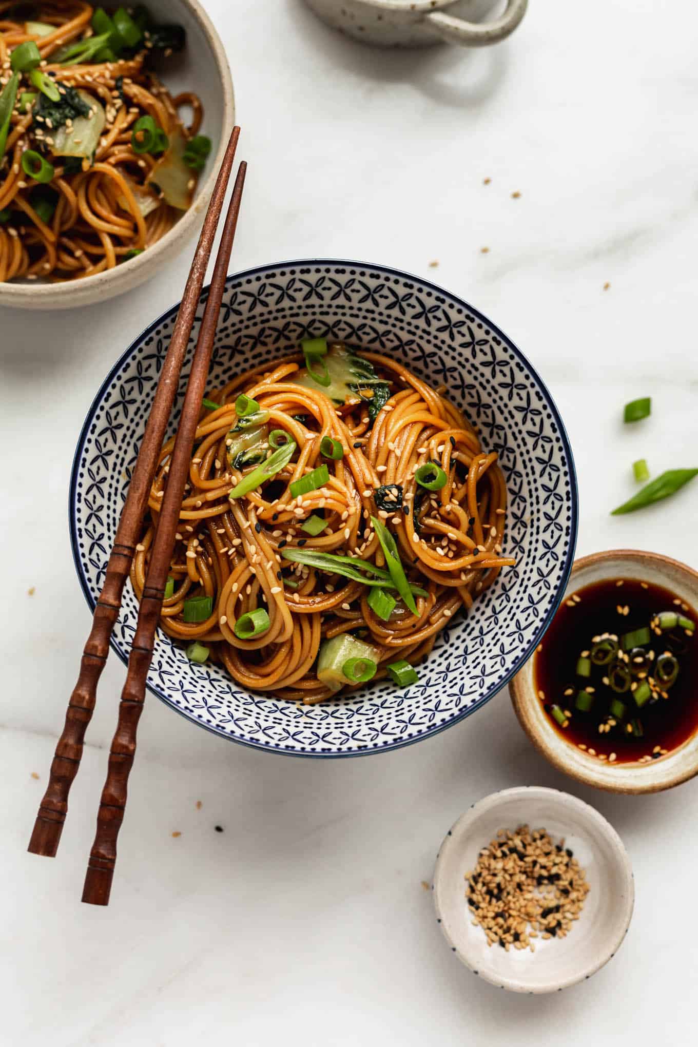 Teriyaki noodles in a blue bowl with a side of teriyaki sauce and sesame seeds