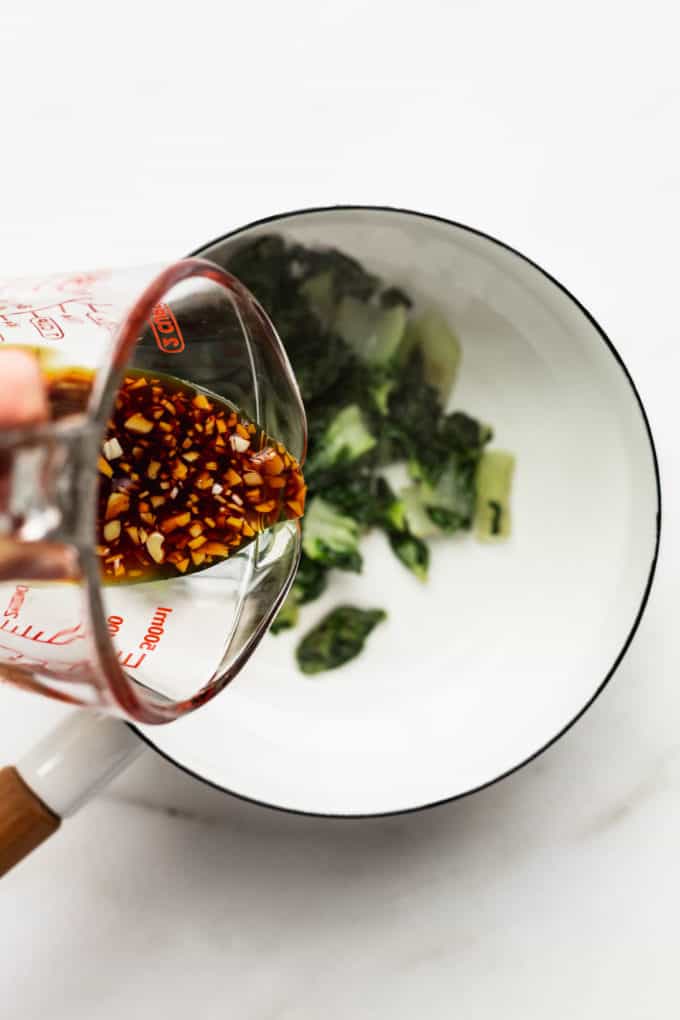 A measuring cup of teriyaki sauce being poured into a white pot of bok choy